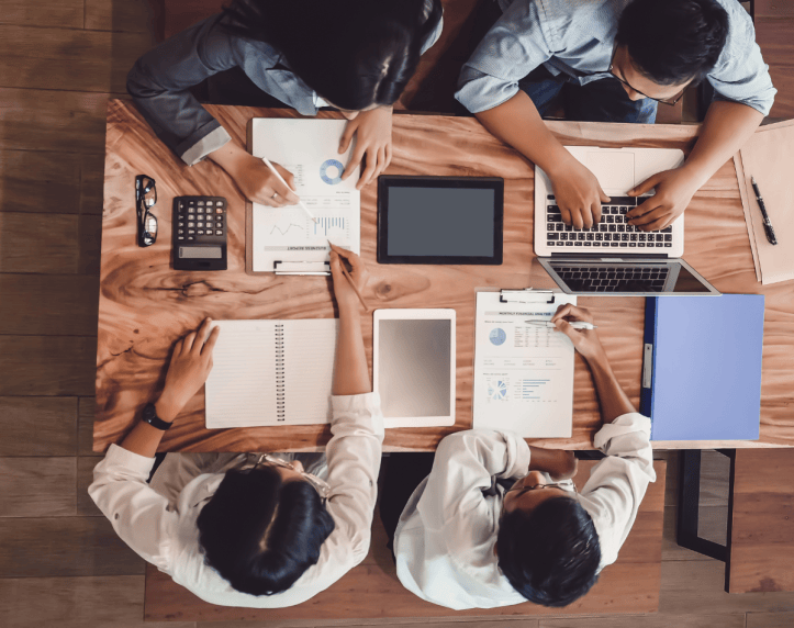 Aerial view of a group of professionals collaborating at a wooden table covered with laptops, tablets, notebooks, and documents with charts and graphs.